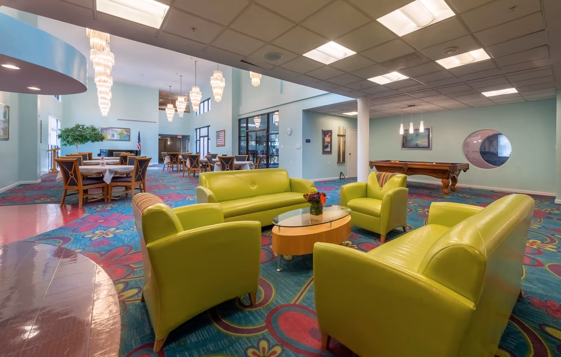 A spacious senior living facility common area with bright green sofas and armchairs arranged around a glass-top coffee table. The room features colorful floral-patterned carpet, multiple dining tables with chairs, a pool table, and large hanging chandeliers. Walls are painted light blue with various framed artworks and a circular window.