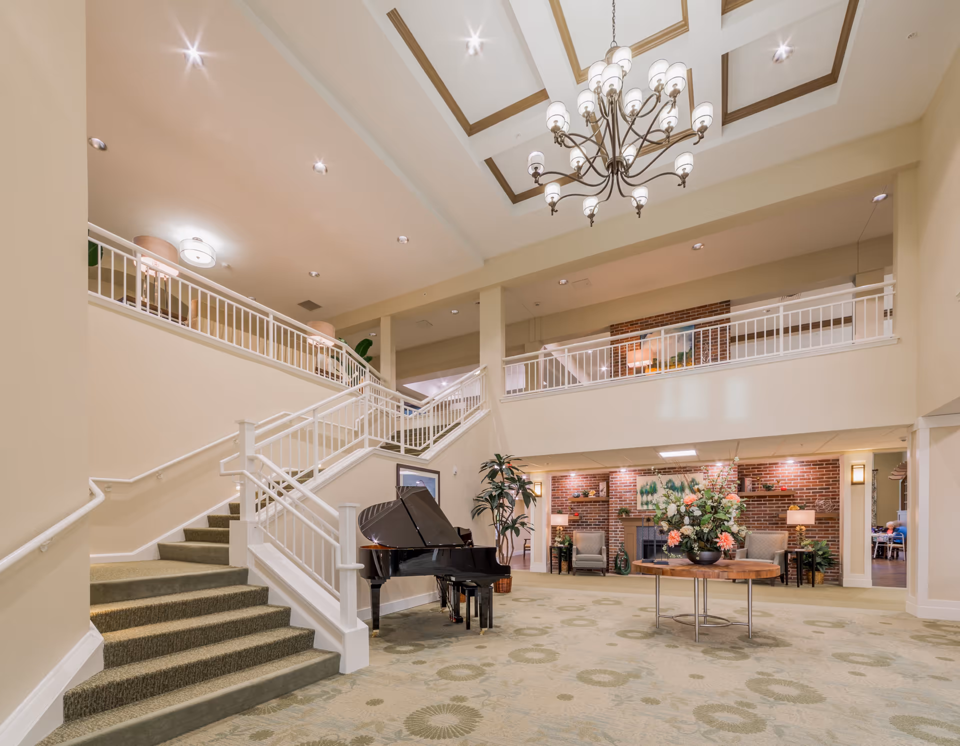 Spacious and well-lit interior of a senior living facility with a grand piano near a staircase leading to an upper level. The area features a large chandelier hanging from a high ceiling, a round table with a floral arrangement, and a cozy seating area with armchairs and lamps against a brick accent wall.