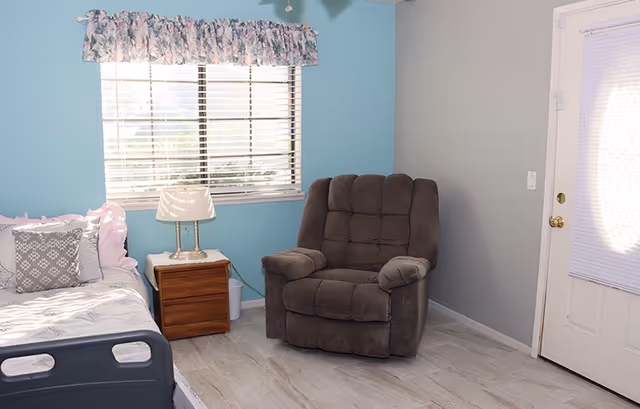 A bedroom with a single bed, wooden nightstand and lamp, a gray recliner, a window with blinds, and a door.