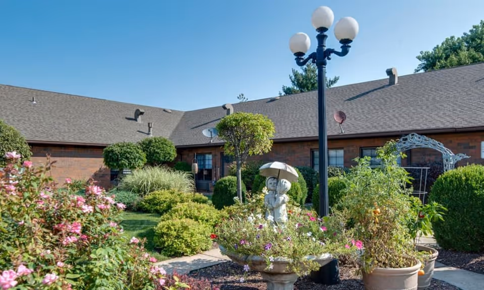 A well-maintained garden area with various green shrubs, flowering plants, and a decorative statue of two children holding an umbrella. There is a black lamppost with three white globes and a brick building with a gray roof in the background under a clear blue sky.