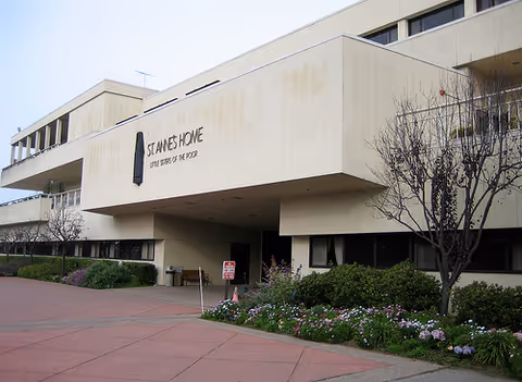 Exterior view of St. Anne's Home For The Aged building showing a beige multi-story structure with the name and motto 'Little Sisters of the Poor' on the front. There are landscaped bushes and flowers along the walkway leading to the entrance.