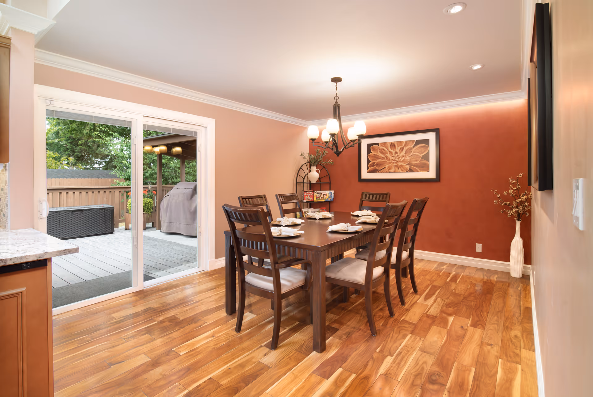 A dining room with a wooden table set for six people, surrounded by six wooden chairs with cushions. The room has hardwood flooring, a red accent wall with a large floral artwork, a chandelier with six lights, and a sliding glass door leading to an outdoor deck area with a grill and seating. There is also a decorative shelf with plants and magazines in the corner.