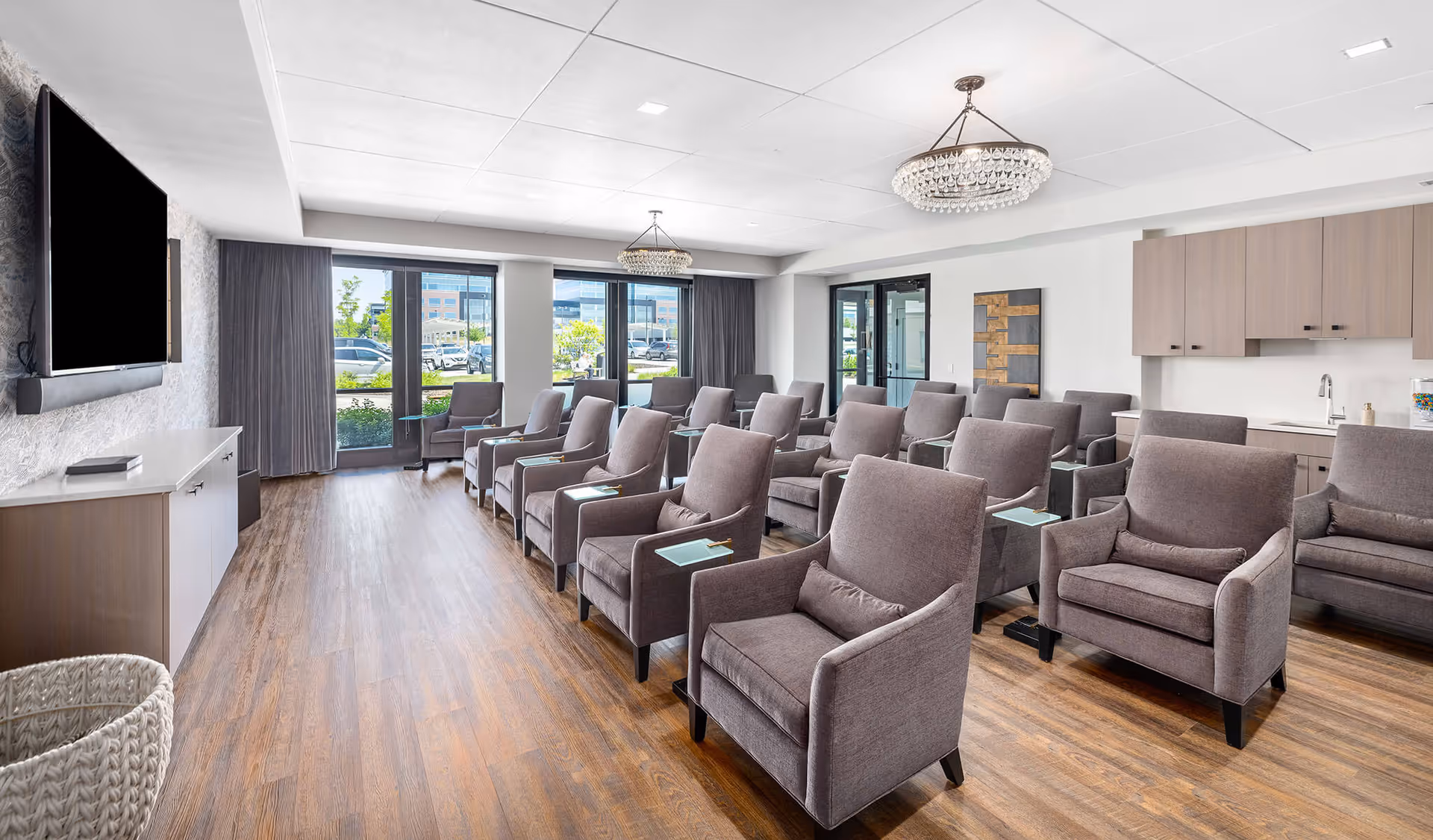 A modern community room with multiple rows of gray upholstered armchairs, each with a small attached side table. The room features wood flooring, two crystal chandeliers, large windows with gray curtains, a wall-mounted flat screen TV, and a kitchenette area with light wood cabinets and a sink.