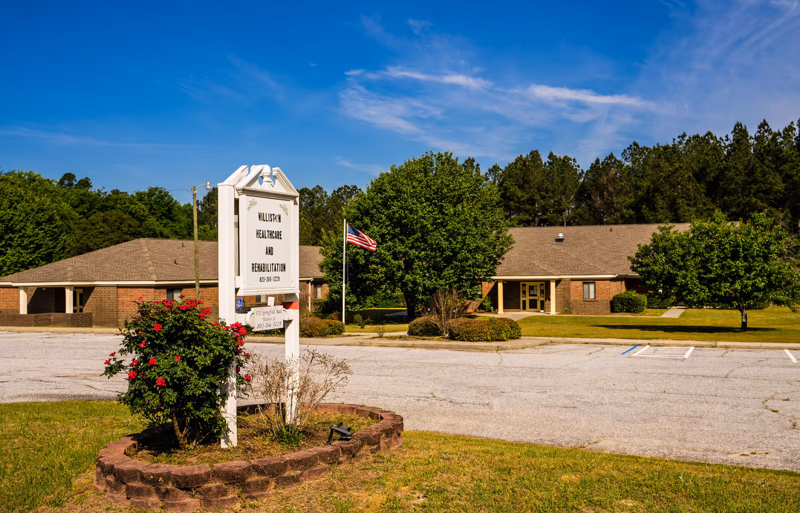 Exterior view of Williston Healthcare and Rehabilitation LLC building with a sign in the foreground surrounded by flowers and greenery, an American flag, and a parking lot under a clear blue sky.