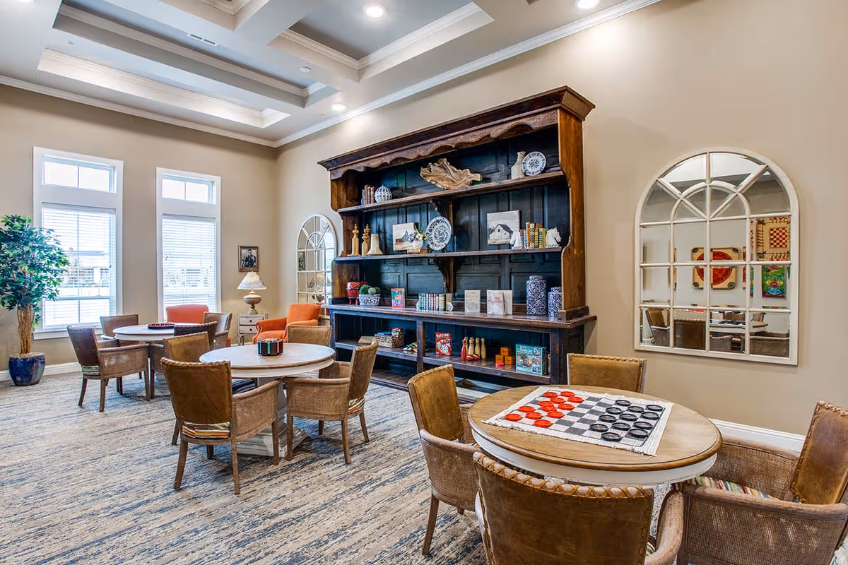 A bright and spacious common area with multiple round tables and chairs arranged for socializing or playing games. One table has a checkers game set up. The room features large windows with blinds, a tall wooden shelving unit filled with decorative items and games, two arched mirrors on the walls, and a potted plant in the corner. The ceiling has recessed lighting and a tray design.