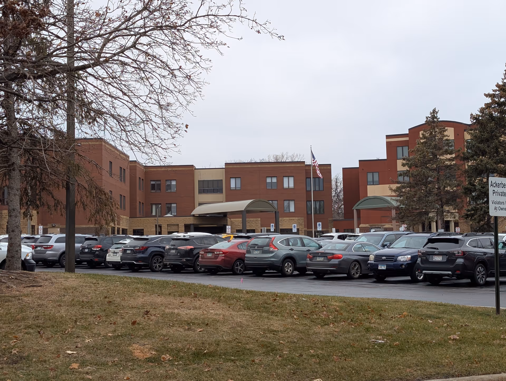 Exterior view of a multi-story brick and tan building with a parking lot full of cars in front. There are two covered entrances and an American flag on a flagpole. Trees with no leaves are visible on the left and right sides of the image under an overcast sky.