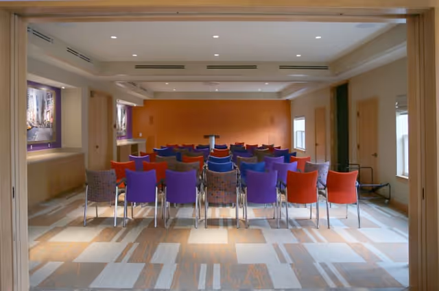 A conference or meeting room with rows of colorful chairs in red, purple, and patterned fabric facing a small podium against an orange accent wall. The room has a modern design with recessed lighting, windows on the right side, and framed artwork on the left wall.