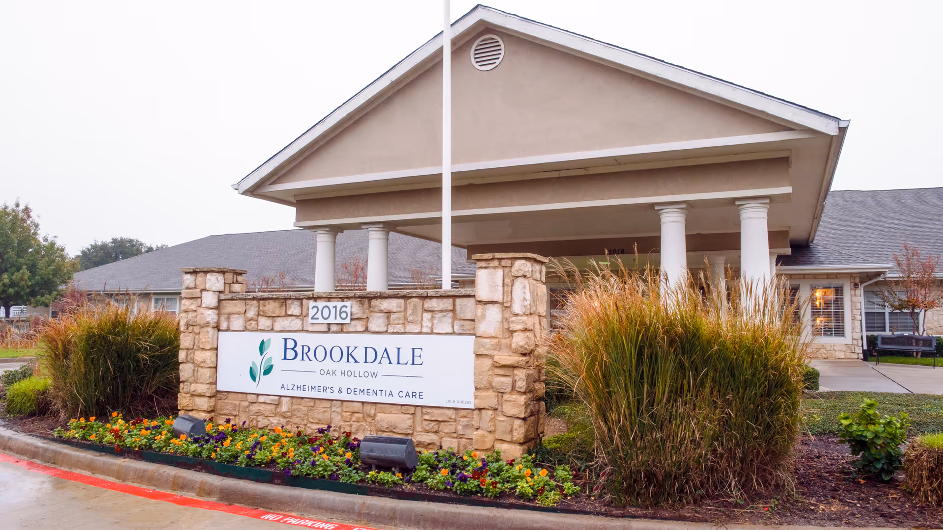 Exterior view of Brookdale Oak Hollow Alzheimer's & Dementia Care facility with a stone sign in front surrounded by flowers and ornamental grasses, and a building with columns and a pitched roof in the background.