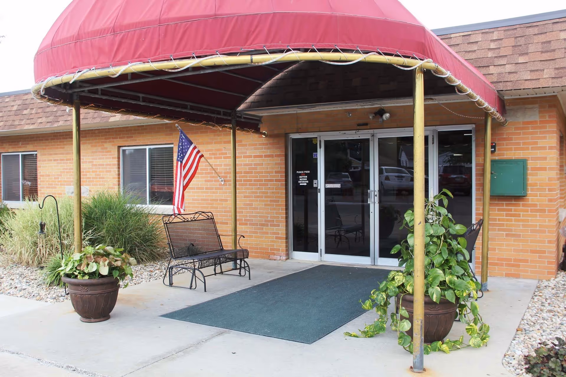 Entrance to a brick building with a red canopy overhead, two potted plants on either side of the entrance, a metal bench with an American flag attached, and double glass doors.