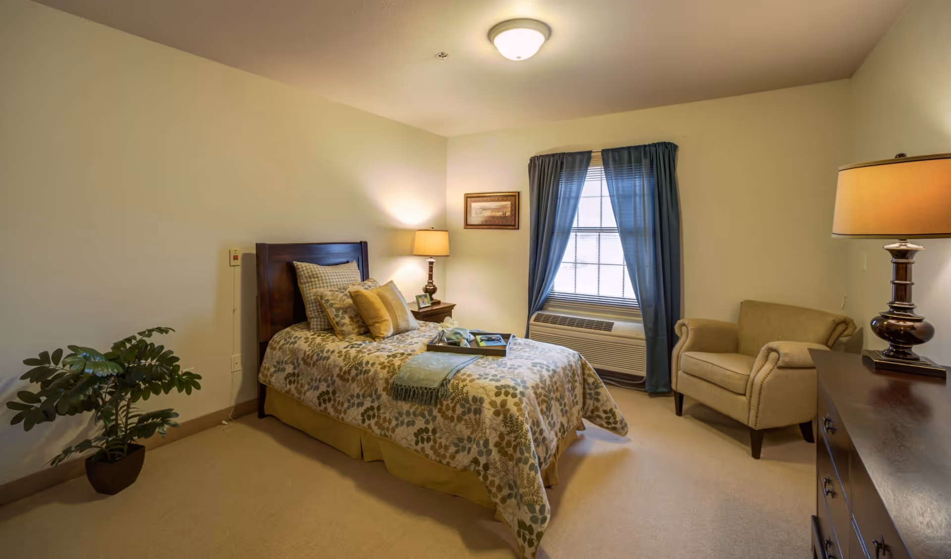 A cozy bedroom in Oak Pointe Assisted Living featuring a single bed with floral bedding and multiple pillows, a wooden nightstand with a lamp and picture frame, a window with blue curtains, a beige armchair, a wooden dresser with a lamp, and a potted plant in the corner.