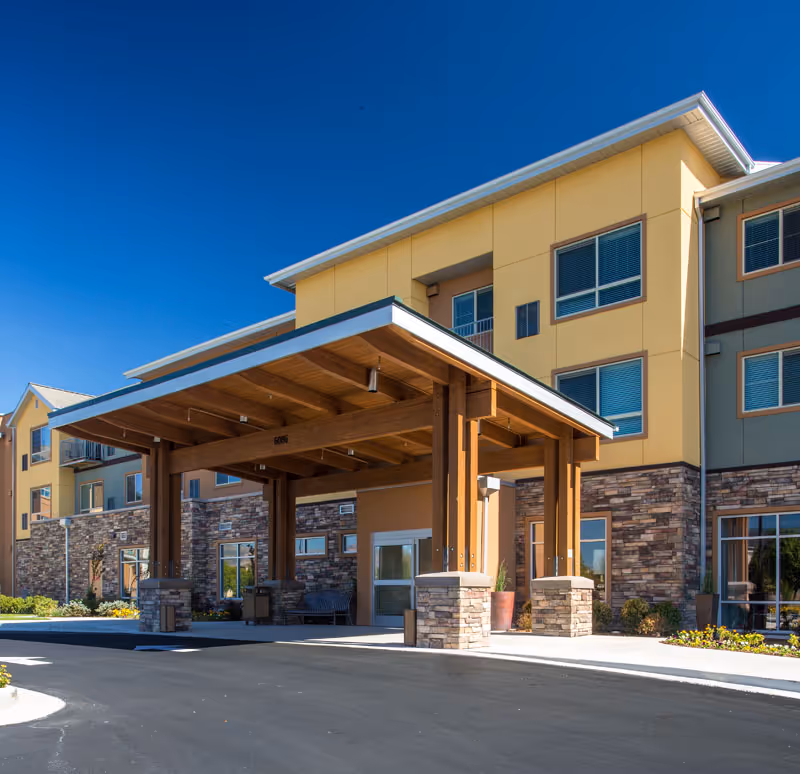 Exterior view of a multi-story senior living facility with a large covered entrance supported by wooden beams and stone pillars. The building features yellow and beige walls with stone accents and multiple windows under a clear blue sky.