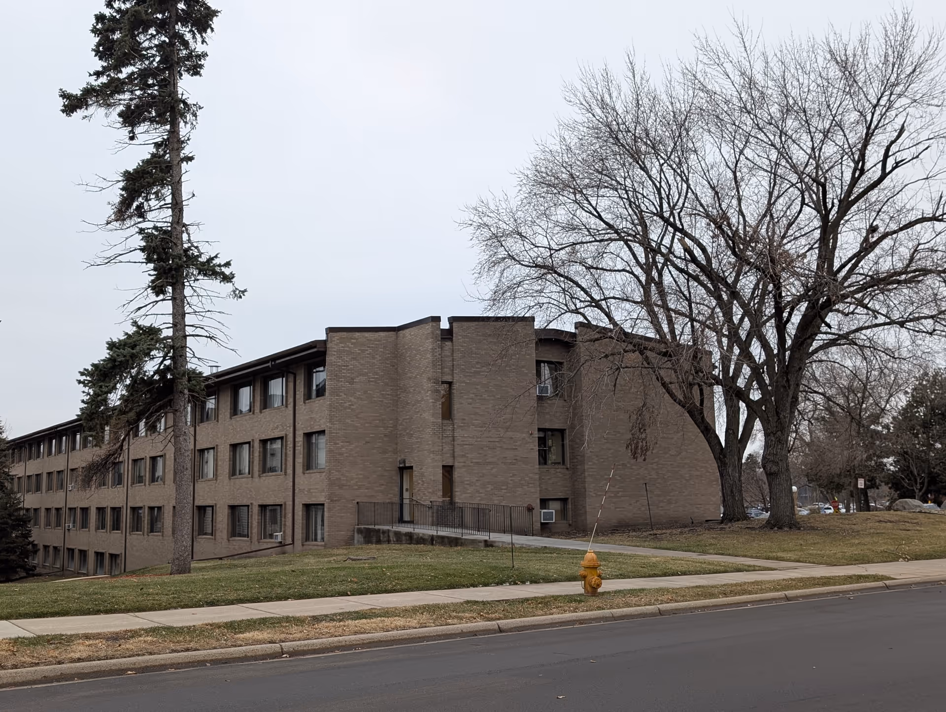 Brick multi-story senior living building with leafless trees and a yellow fire hydrant on the grassy front lawn.