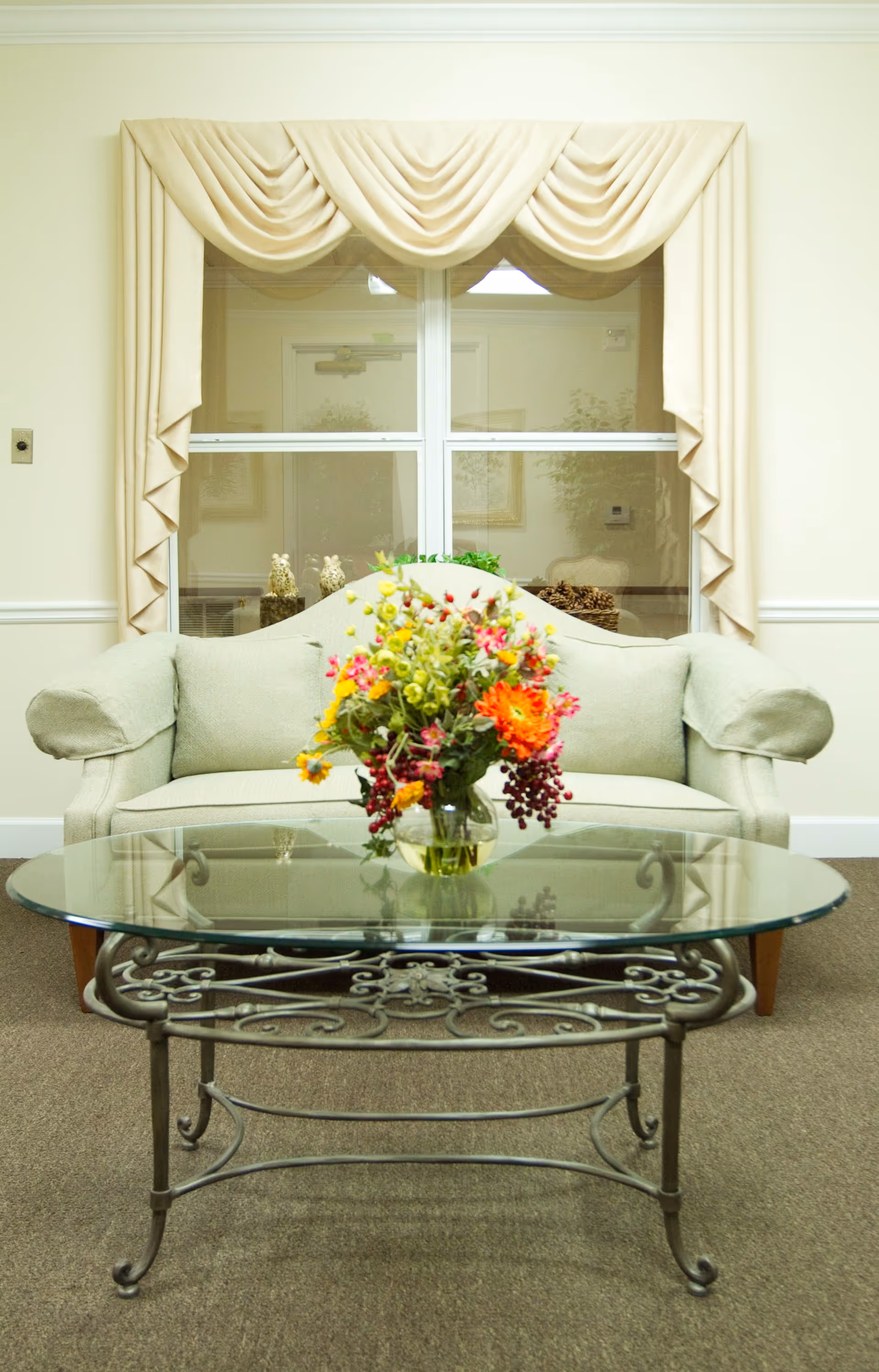 A cozy living room area with a light green upholstered sofa placed against a wall with a window. The window is adorned with cream-colored draped curtains. In front of the sofa is a glass-top coffee table with an ornate metal frame, holding a vase filled with a colorful bouquet of flowers.