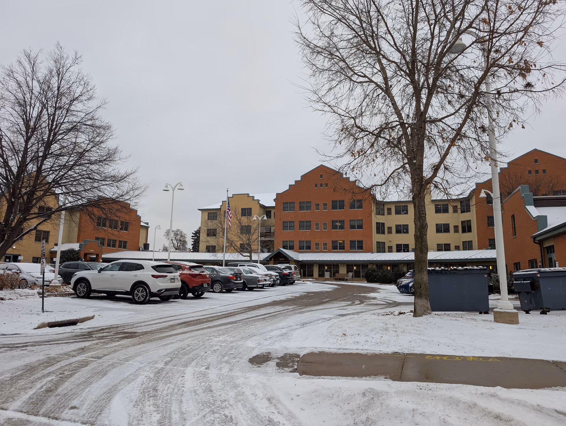 Snow-dusted parking lot and driveway leading to the front entrance of a multi-story senior living building with parked cars and bare trees.