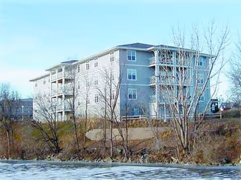 A four-story residential building with multiple balconies, situated near a body of water with leafless trees in the foreground under a clear blue sky.