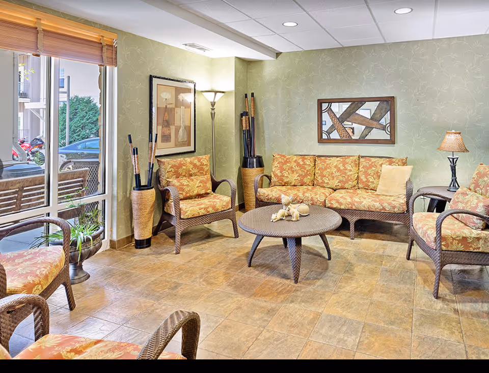 Sunlit common lounge with wicker sofa and matching chairs arranged around a round coffee table, artwork on the walls, and a floor lamp.