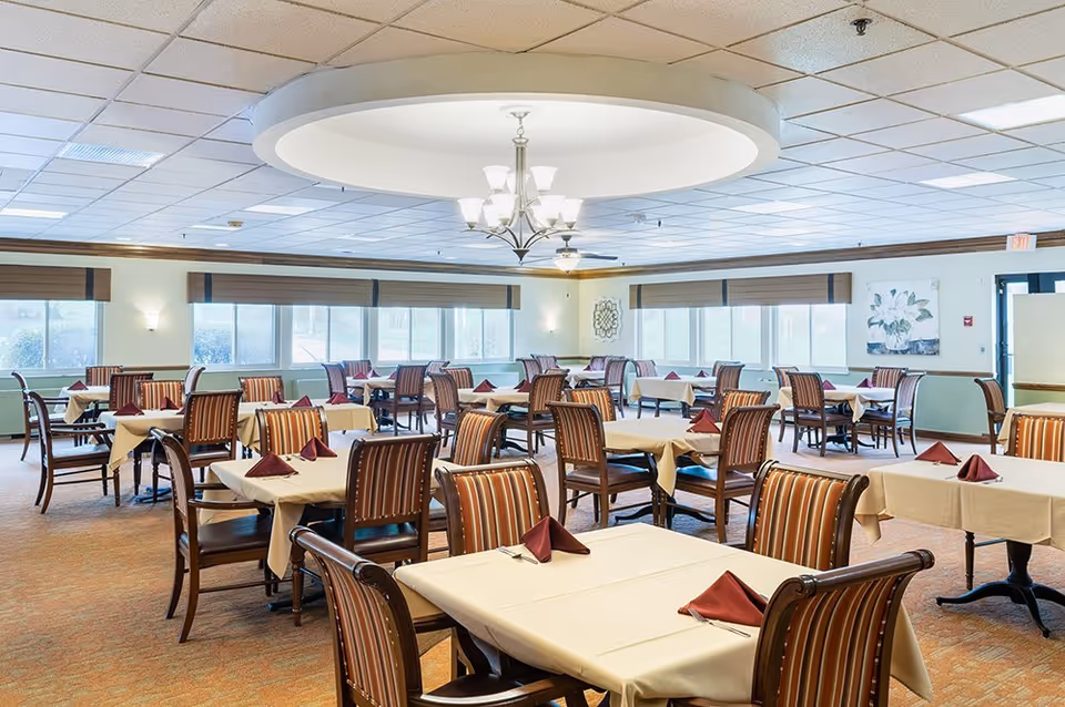 A spacious dining room with multiple tables covered in beige tablecloths and set with folded burgundy napkins. The room has large windows with brown blinds, a beige carpet, and a ceiling light fixture with a circular recessed design.