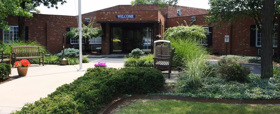 Front exterior view of a single-story brick building with a 'WELCOME' sign above the entrance. The entrance is surrounded by greenery, including bushes, potted flowers, and trees. There are benches and a trash can near the walkway leading to the door.