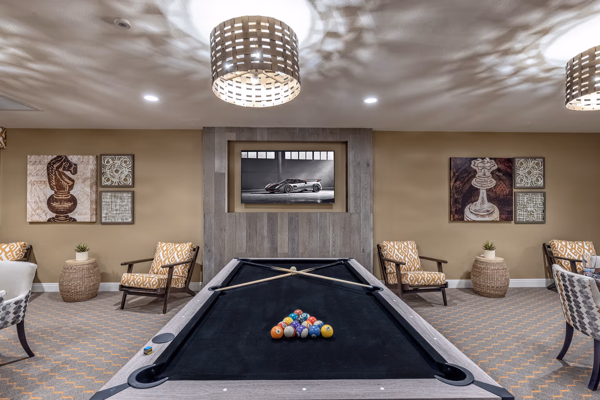 A recreational room with a pool table in the center, two pool cues crossed on the table, and a set of billiard balls arranged for a game. The room has patterned carpet, beige walls adorned with chess-themed artwork, and several cushioned chairs with patterned upholstery. A modern light fixture hangs from the ceiling, casting decorative shadows. A framed picture of a sports car is mounted on a wooden accent wall behind the pool table.