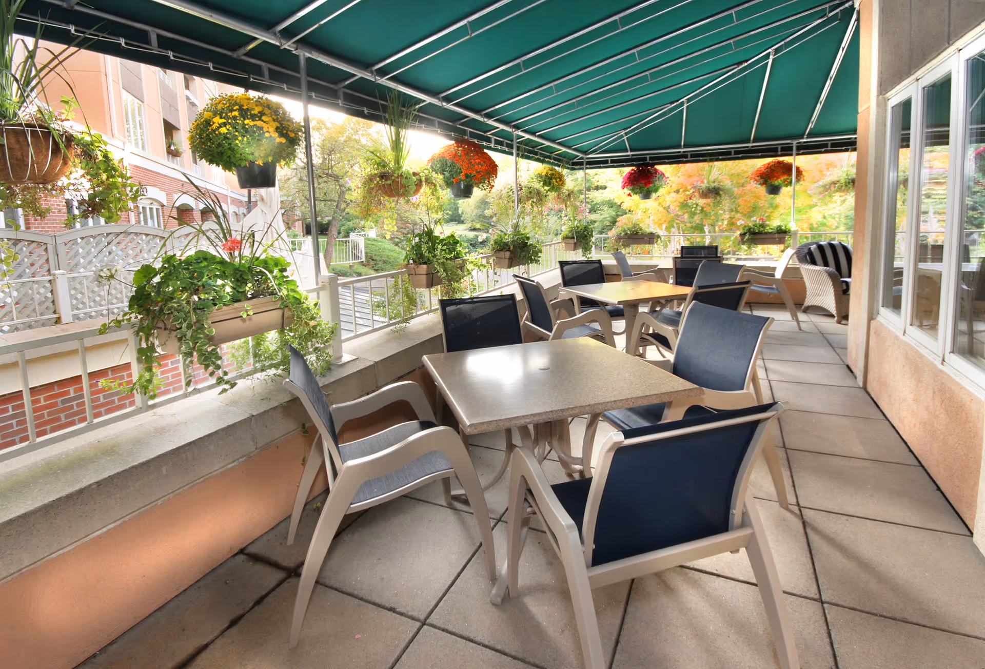Outdoor covered patio area with multiple tables and chairs arranged for seating. Hanging flower pots with colorful flowers and greenery decorate the space. The patio is adjacent to a building with windows and overlooks a garden area with trees and shrubs.