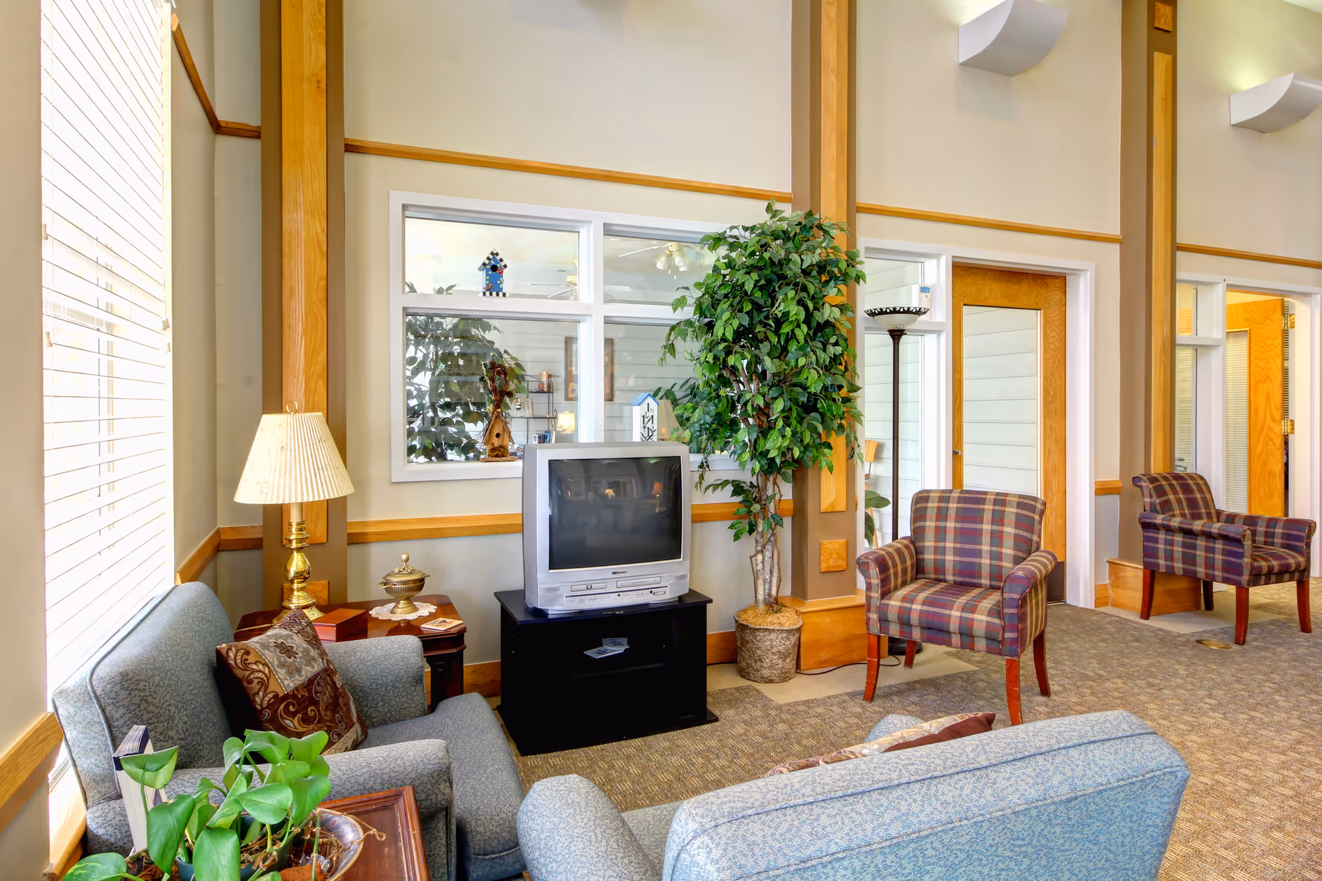A cozy living room area with patterned armchairs and a blue sofa arranged around a small black TV stand holding an older model television. There is a tall potted plant beside the TV, a side table with a lamp and decorative items, and large windows with white blinds letting in natural light. The walls are beige with wooden trim accents, and there are doors and windows leading to other rooms in the background.