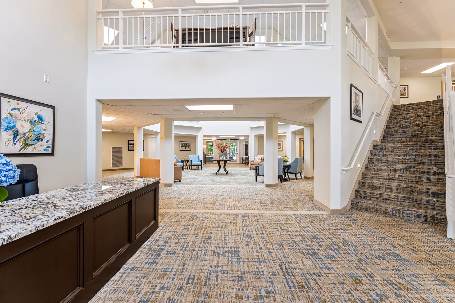 Spacious, well-lit senior living facility lobby with a reception desk, seating areas, and a carpeted staircase.