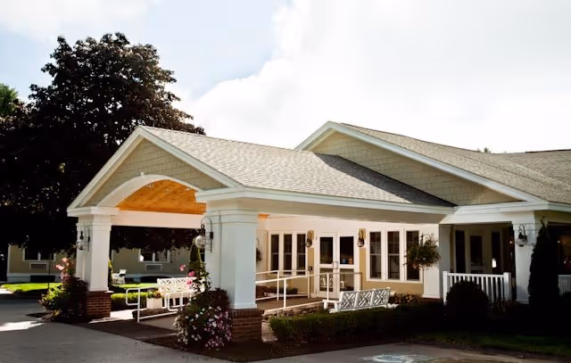 Exterior view of the entrance to a senior living facility with a covered drop-off area, white columns, hanging flower baskets, benches, and a ramp leading to glass doors. Trees and a clear sky are visible in the background.