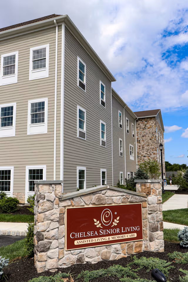 Stone entrance sign reading "Chelsea Senior Living" in front of a multi-story senior living building under a partly cloudy sky.