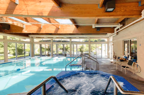 Bright indoor swimming pool and bubbling hot tub in a sunlit room with large windows and exposed wooden beams.