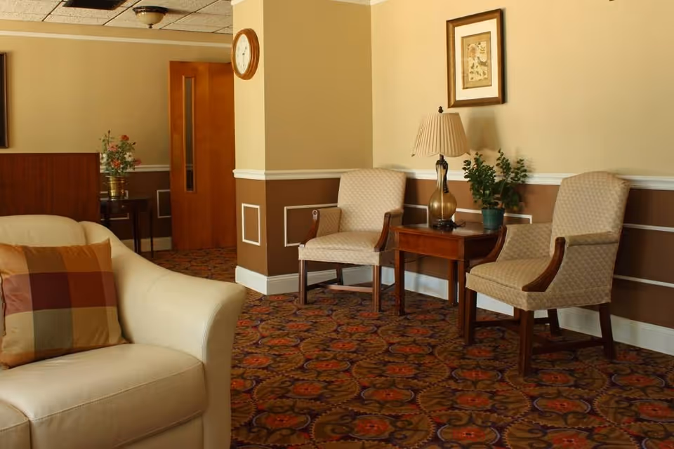 A cozy seating area in a senior living facility with two beige upholstered chairs, a wooden side table with a lamp and a potted plant, a beige couch with a multicolored plaid pillow, a wall clock, and framed artwork on the wall. The carpet has a floral pattern in warm colors.