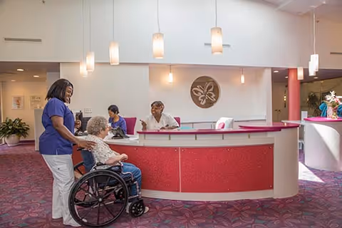 A senior woman in a wheelchair is being pushed by a caregiver in a blue uniform inside a facility. Two other staff members are seated behind a curved reception desk with a pink front panel. The area has purple patterned carpet, pendant lights hanging from the ceiling, and a decorative wall emblem behind the desk.