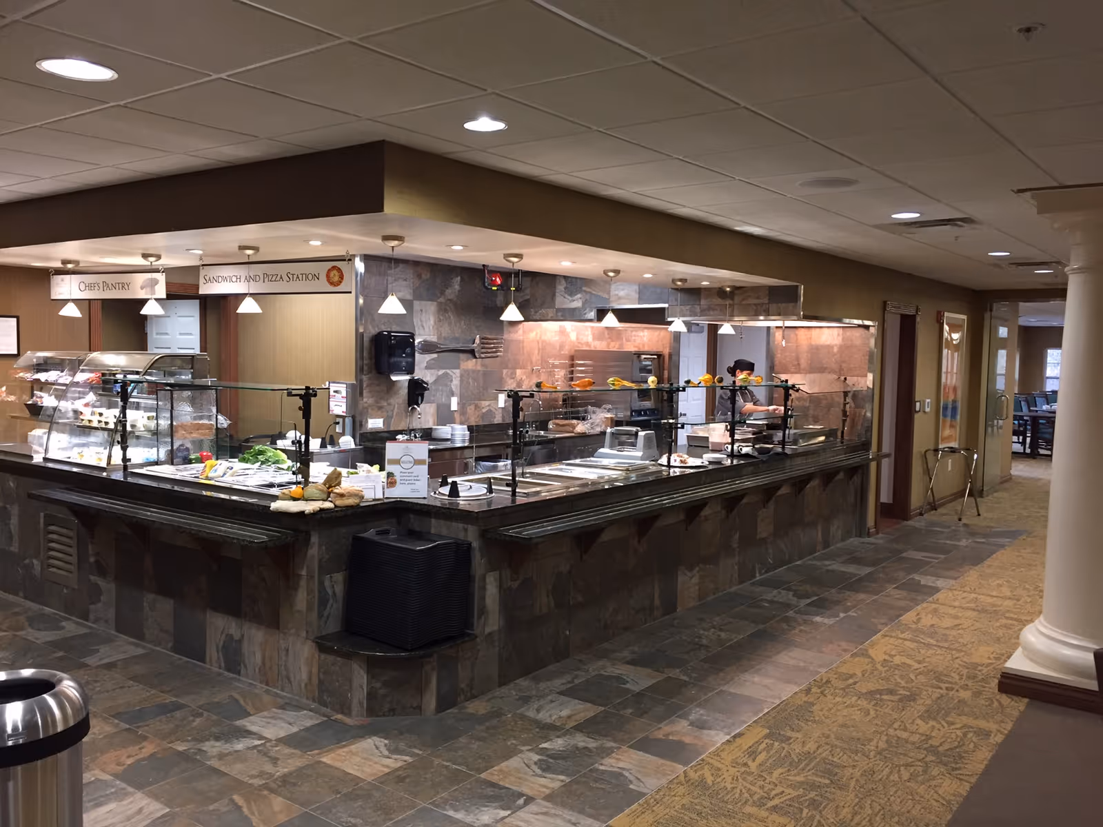 Cafeteria-style serving counter with glass guards and food displays in a retirement facility dining area.