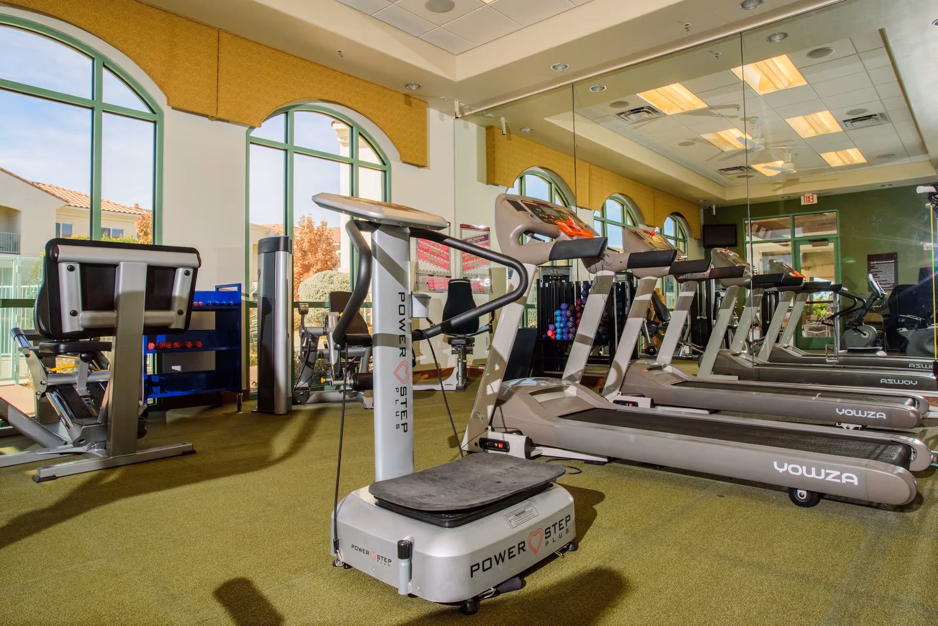 Bright fitness room with treadmills, exercise machines and a PowerStep platform in front of large arched windows and a mirrored wall.