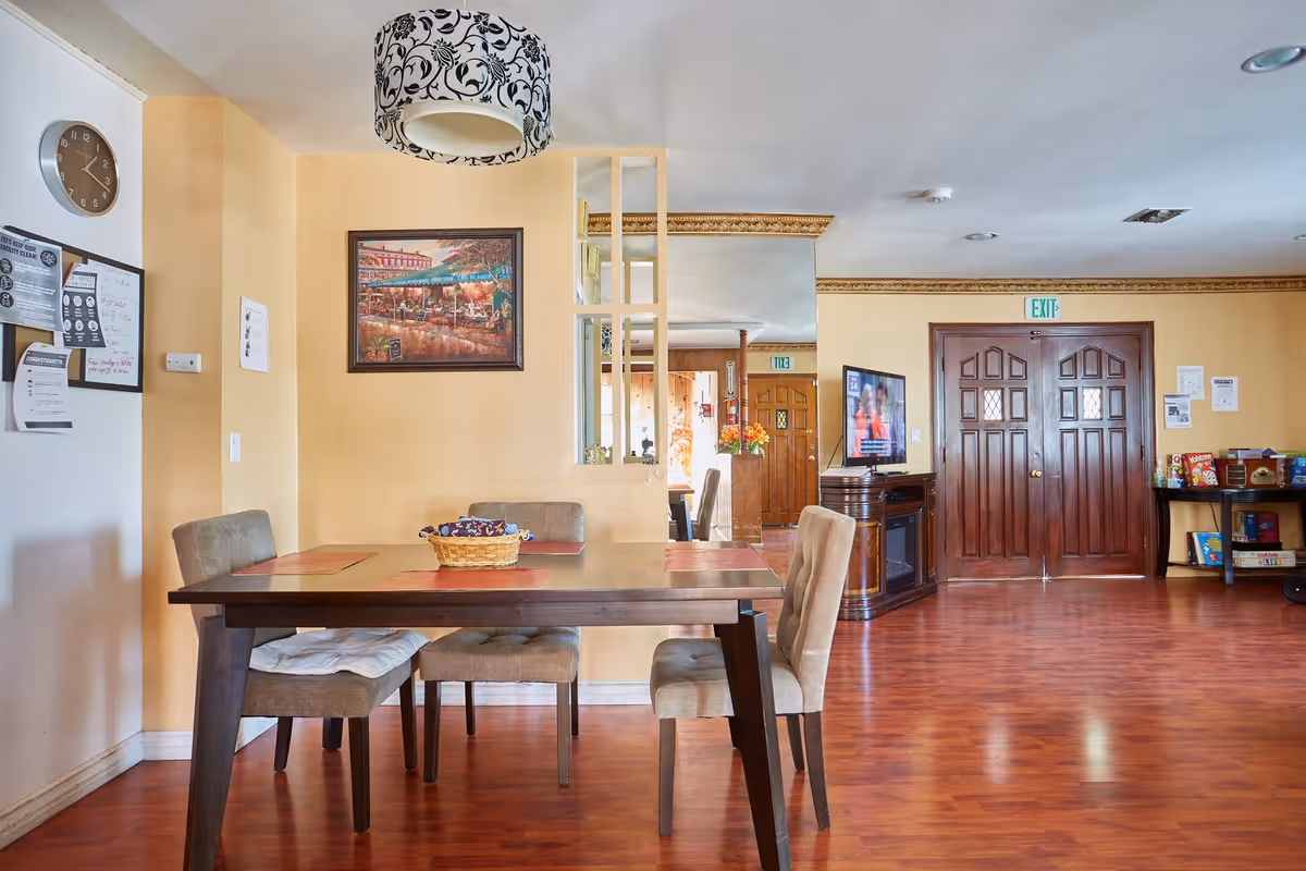 Interior view of a senior living facility dining area with a wooden table and four cushioned chairs. The walls are painted yellow, and there is a decorative hanging lamp above the table. A framed painting hangs on the wall, and a large mirror with vertical wooden slats is mounted next to it. In the background, there is a wooden double door with an exit sign above it, a TV on a stand, and a small table with board games and snacks. The floor is a polished wood finish.