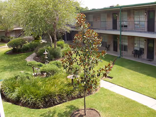 Outdoor courtyard area with green grass, landscaped garden beds, small trees, and a two-story building with balconies and patio chairs.