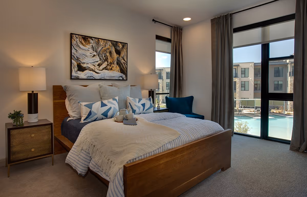 A cozy bedroom with a wooden bed frame, neatly made bed with multiple pillows and a beige throw blanket. There are two bedside tables with modern lamps and a small plant on one side. A large window with curtains offers a view of an outdoor pool and adjacent buildings. A blue armchair is placed near the window, and a framed abstract artwork hangs above the bed.