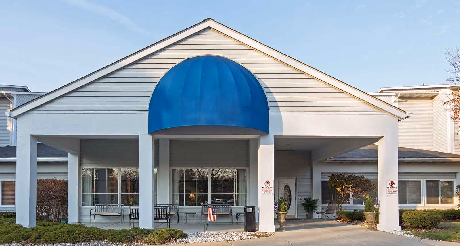 Front exterior view of a senior living facility named Sterling Place with a white building, large windows, a blue awning over the entrance, benches on the porch, and landscaping with bushes and small trees.