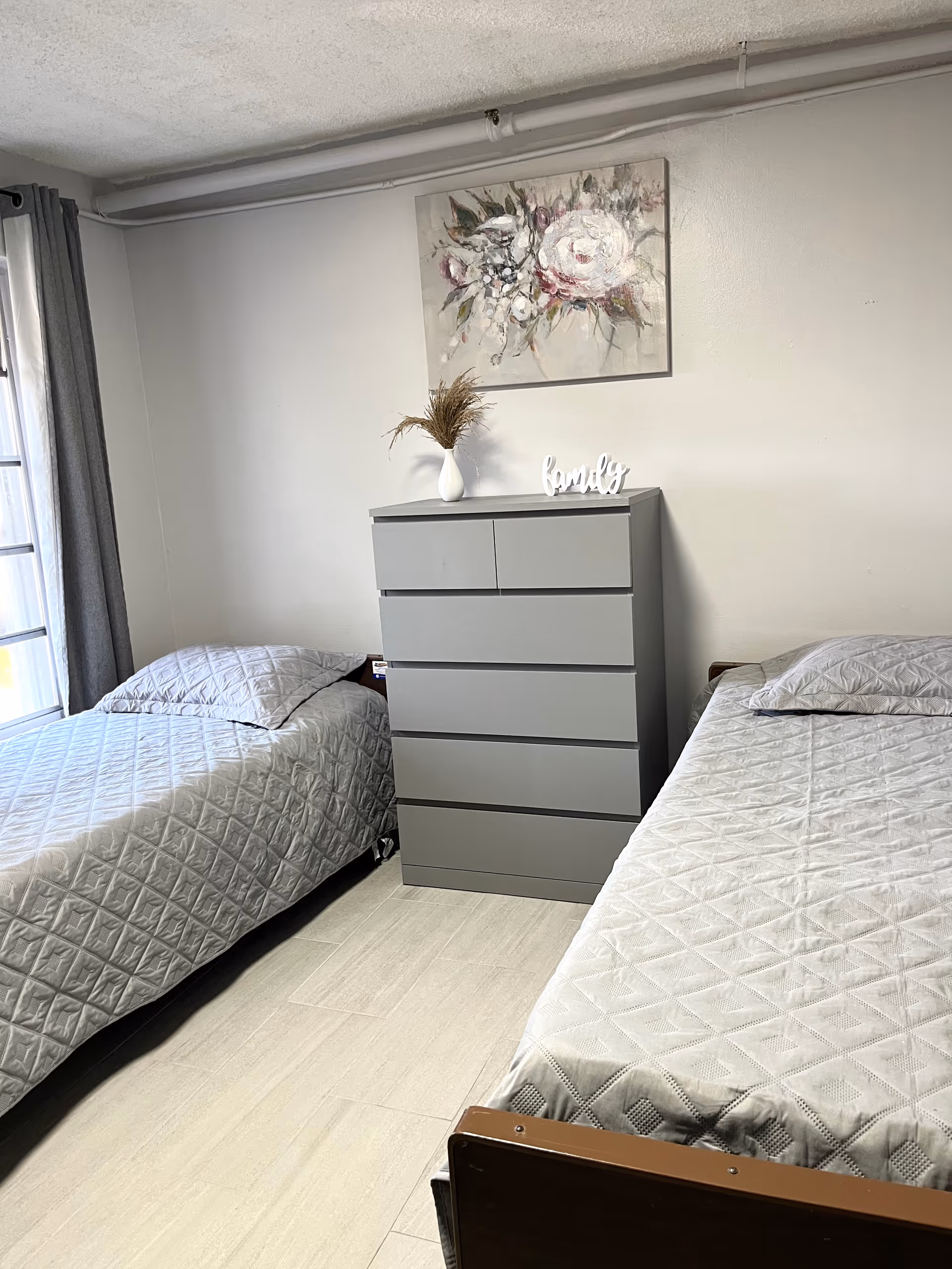 A simple bedroom with two single beds covered in light gray quilted bedspreads. Between the beds is a gray chest of drawers with a white vase holding dried plants and a decorative word 'family' on top. Above the chest hangs a floral painting. A window with gray curtains is visible on the left side.