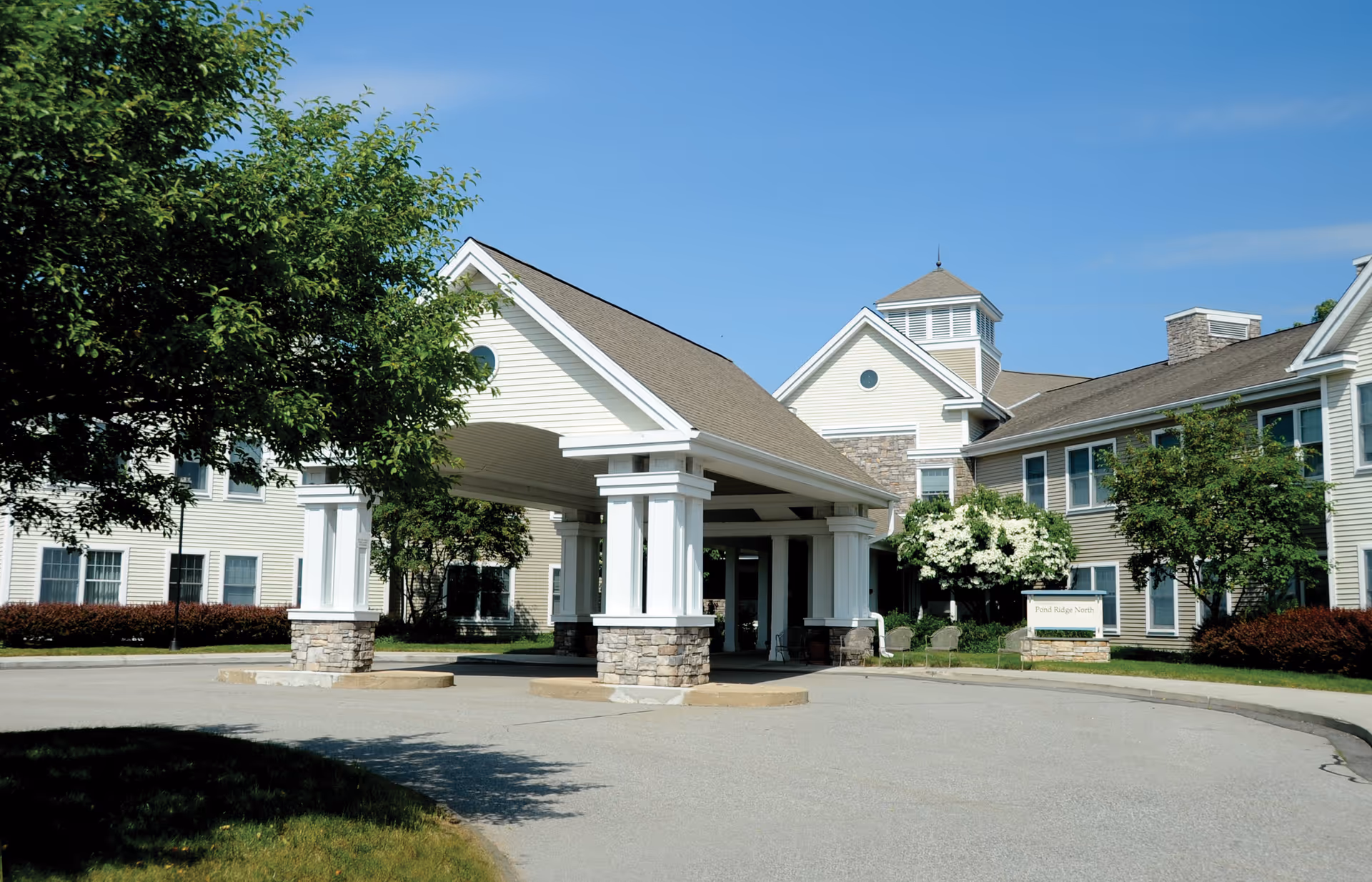 Exterior view of a senior living facility building with a covered entrance supported by white columns with stone bases. The building is light-colored with multiple windows and surrounded by trees and shrubs under a clear blue sky.