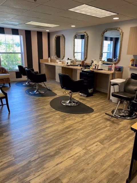 Interior view of a salon area with three black styling chairs in front of a counter with three ornate mirrors mounted on the wall. The room has wood flooring, a large window with striped curtains, and various hair styling tools and products on the counter.