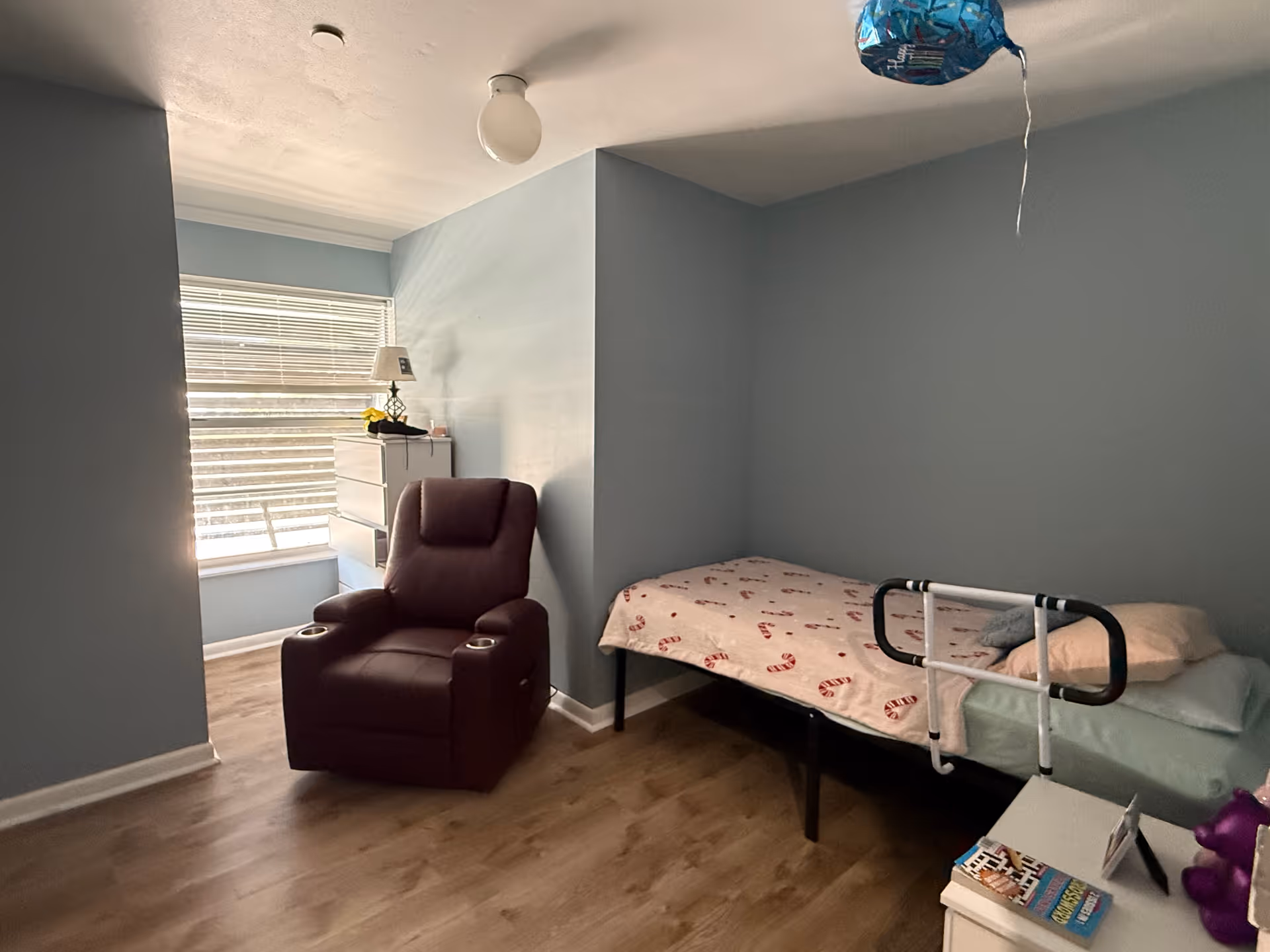 A small bedroom with light blue walls and wood flooring featuring a single bed with a candy cane patterned blanket and a safety bed rail. Next to the bed is a white nightstand with books and a purple teddy bear. There is a maroon recliner chair near a window with blinds, and a white dresser with a lamp and some items on top. A blue balloon is floating near the ceiling.