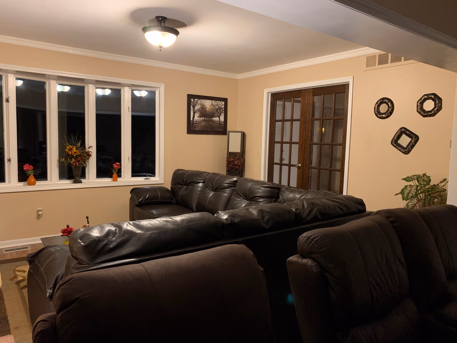 A cozy living room with beige walls, a ceiling light fixture, and a large window with white trim. The room features a dark brown leather sectional sofa and a matching recliner. On the wall, there is a framed picture of a tree-lined path and three decorative wall hangings. A tall mirror and a potted plant are placed in the corner near wooden French doors.