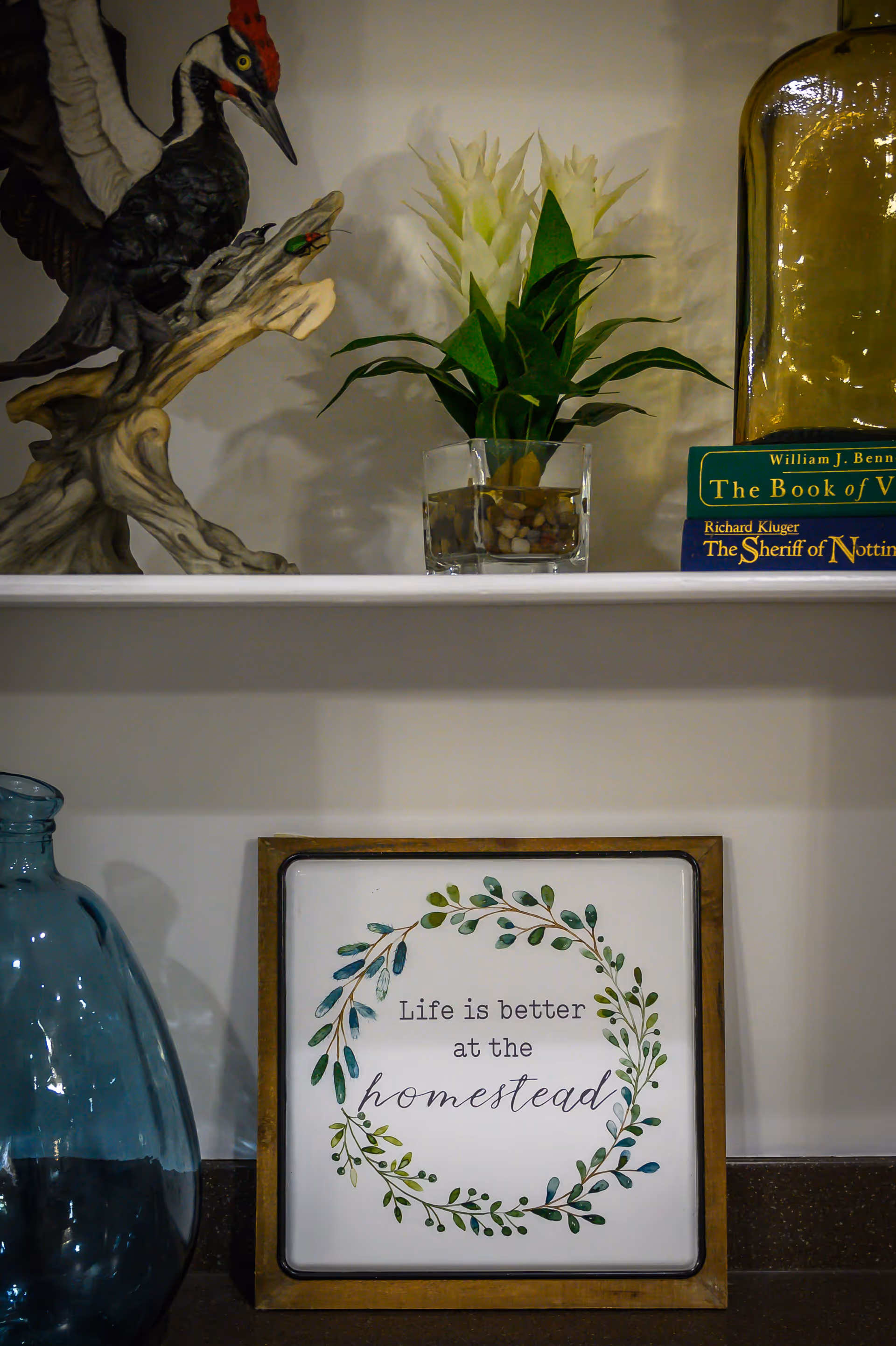A decorative shelf with a wooden bird sculpture, a small plant in a glass container, two stacked books, a large glass bottle, a blue glass vase, and a framed sign that reads 'Life is better at the homestead' surrounded by a leafy wreath design.