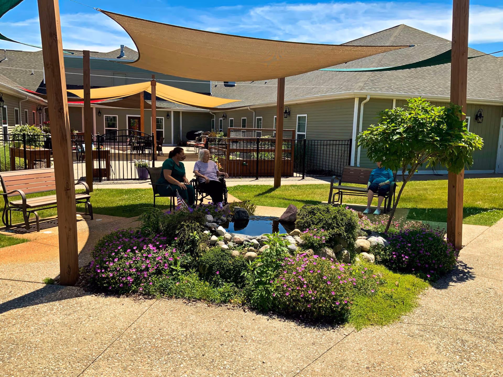 Outdoor courtyard area at Vicinia Gardens Luxury Retirement Living with shaded seating areas, benches, a small pond surrounded by flowers and greenery, and two elderly women sitting and conversing, one in a wheelchair and another seated on a chair, with a third person sitting on a bench nearby.