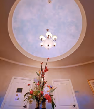 View looking up at a circular ceiling with a chandelier in the center, above a floral arrangement in a vase. Two white doors are visible on the walls below the ceiling.