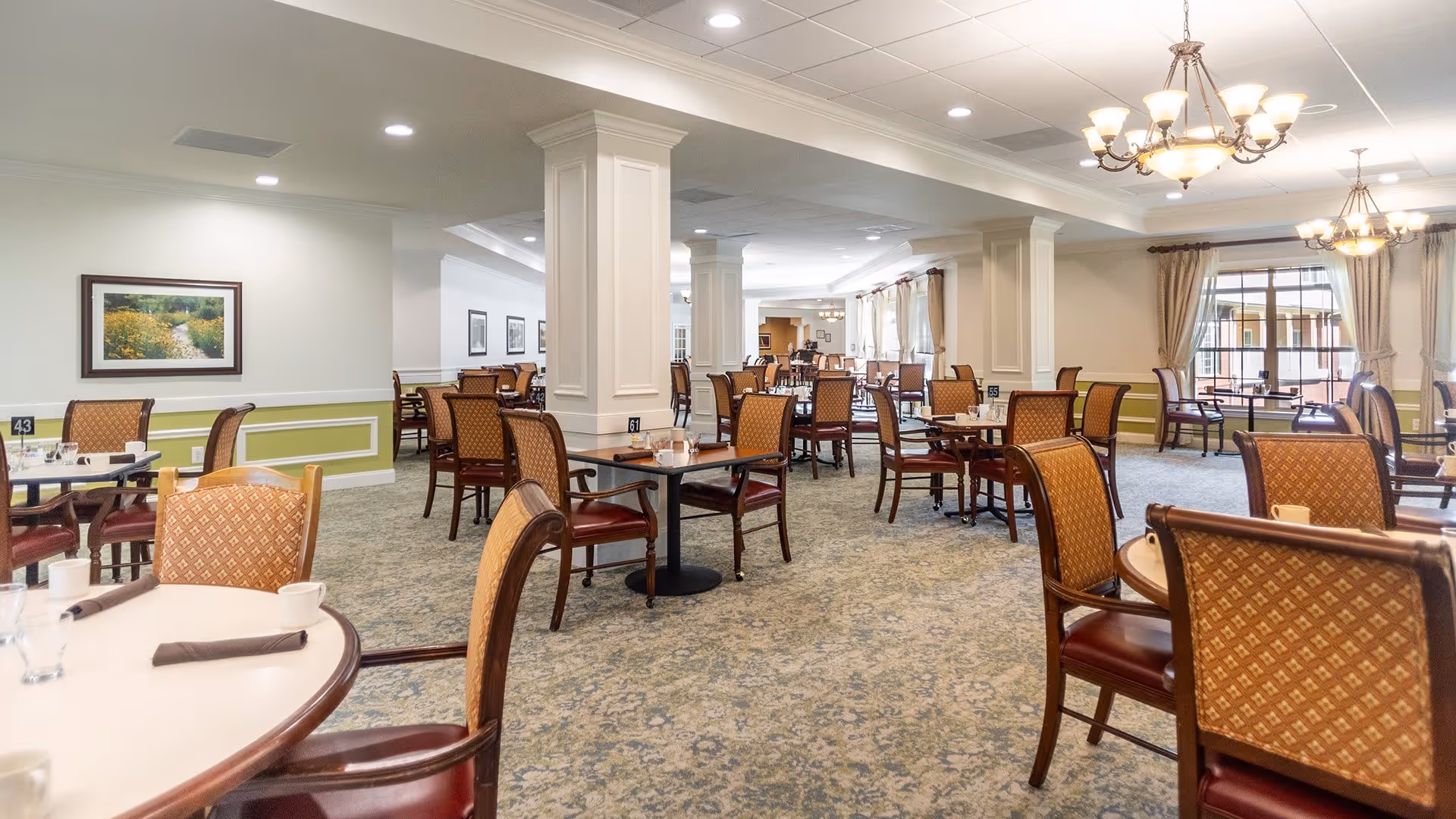A spacious dining room with multiple round tables and cushioned chairs arranged neatly. The room features large windows with curtains, chandeliers hanging from the ceiling, and framed artwork on the walls. The carpet has a patterned design, and the overall ambiance is bright and inviting.