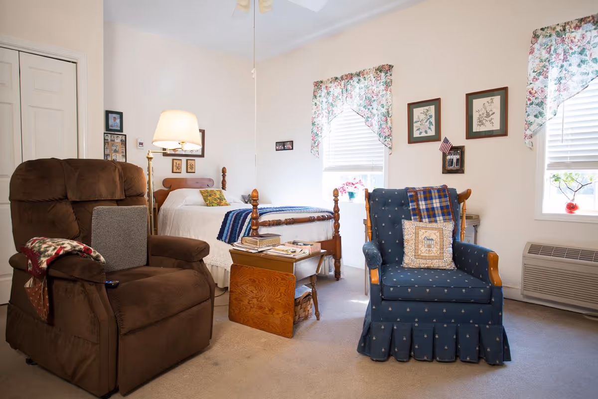 A cozy bedroom with a wooden bed covered in a white bedspread and a blue striped blanket. There is a brown recliner chair with a gray cushion and a patterned throw, and a blue upholstered armchair with a decorative pillow and a plaid blanket. The room has two windows with floral curtains, framed pictures on the walls, a wooden side table with books, and a floor lamp beside the bed.