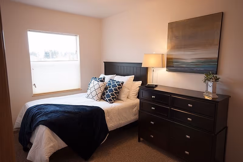 A cozy bedroom with a neatly made bed featuring white bedding, a dark blue throw blanket, and decorative pillows in white and blue patterns. Next to the bed is a dark wooden dresser with a lamp, a small plant, and a decorative clock on top. A large abstract painting hangs on the wall above the dresser. A window with a partially closed blind allows natural light into the room.