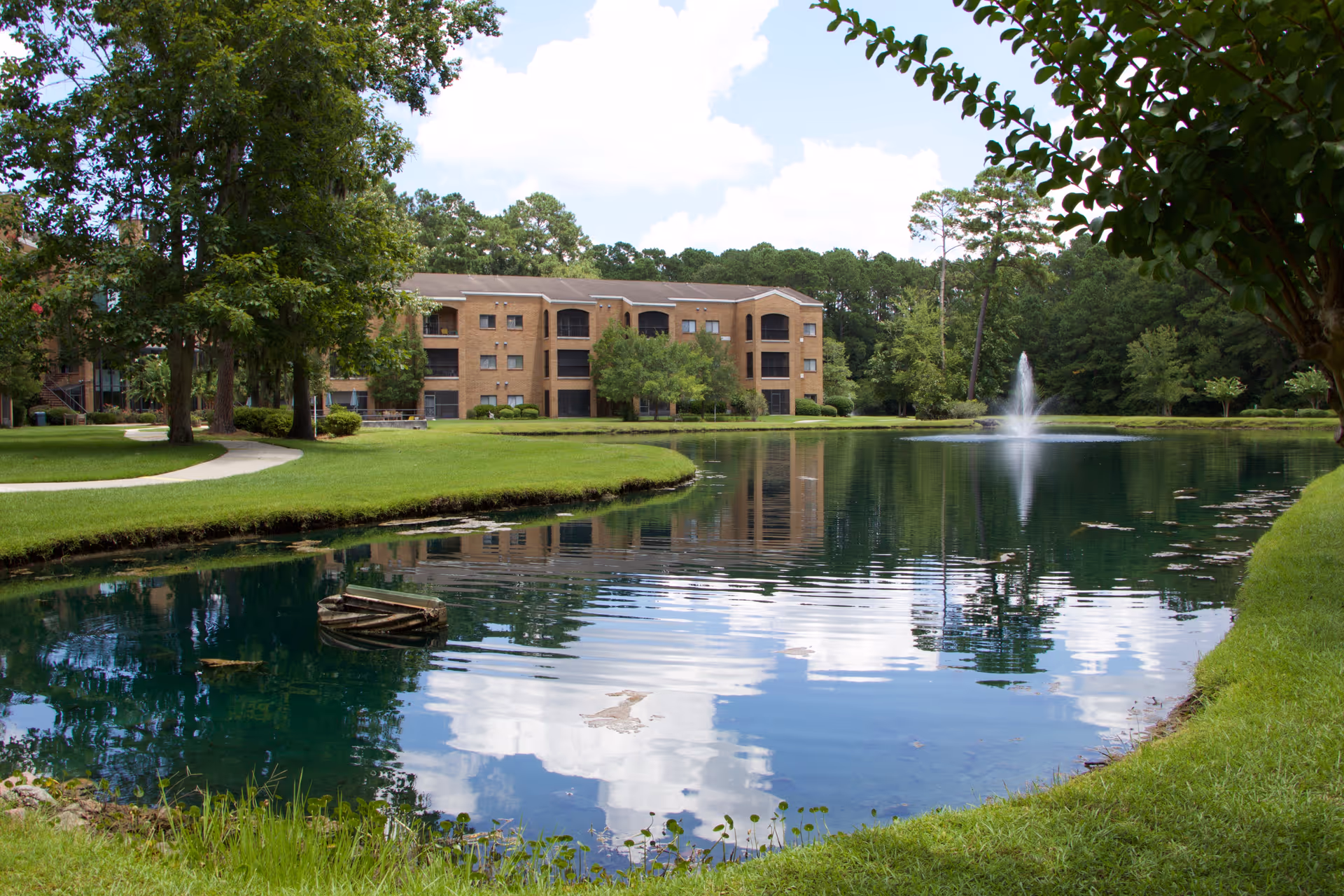 Brick residential building seen across a pond with a fountain, surrounded by green lawns and trees.