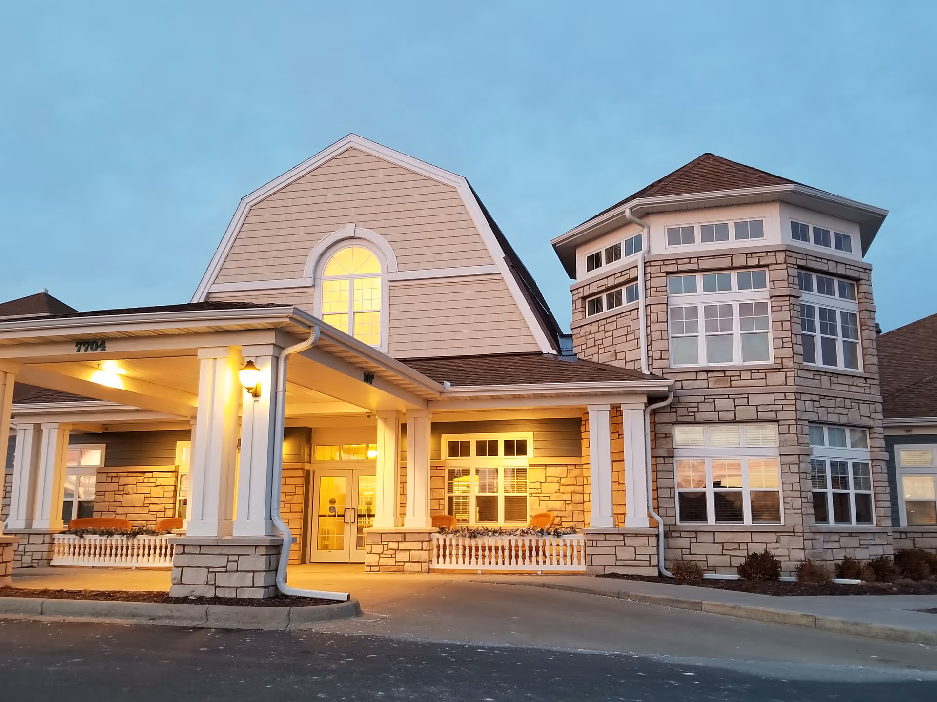 Front entrance of a multi-story memory care building with columns, a covered drive-up, and warm interior lights at dusk.